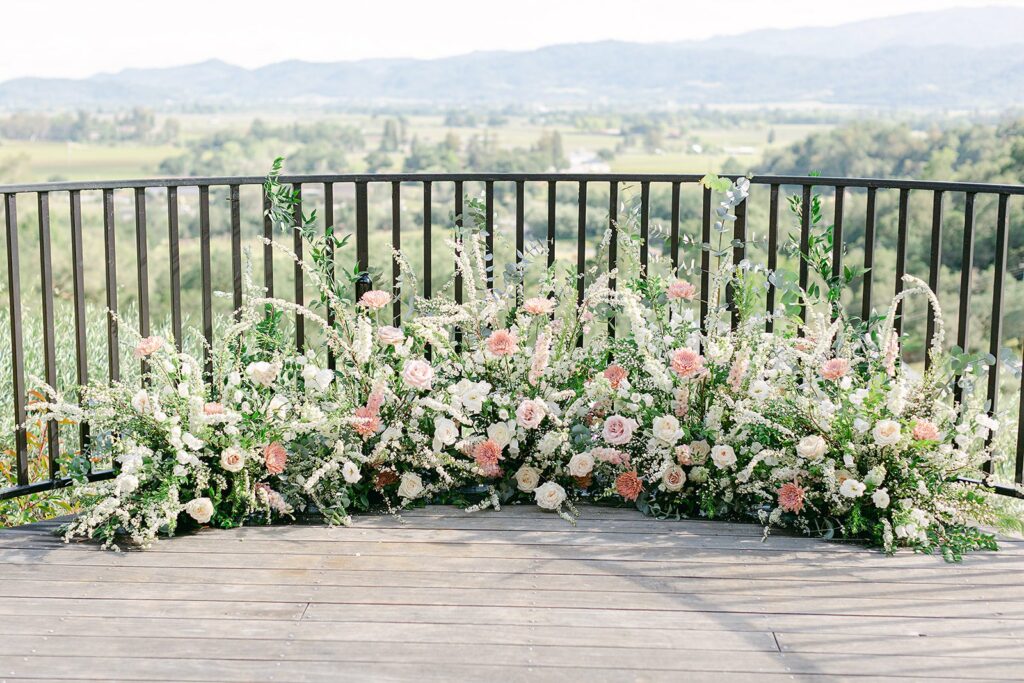 A lush floral arrangement with various pink, white, and green flowers is placed on a wooden deck with a black metal railing, overlooking a scenic valley and hills in the background.