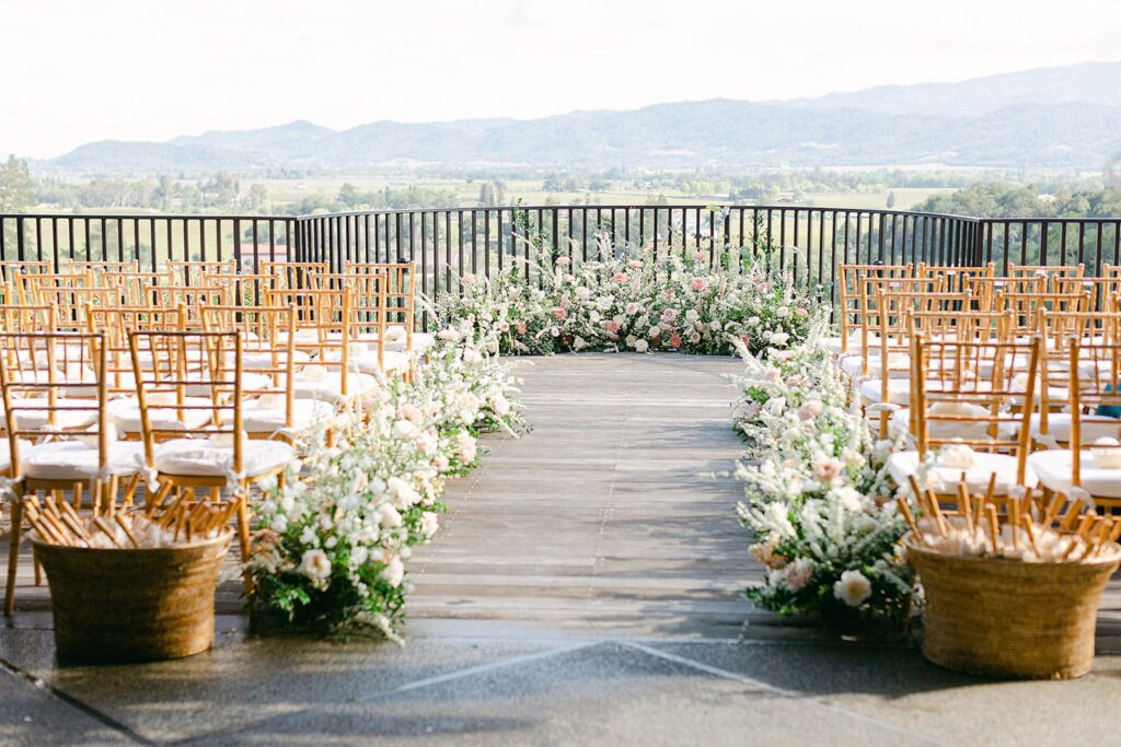 A wooden outdoor wedding ceremony setup with rows of wooden chairs adorned with white cushions, floral arrangements along the aisle, and a scenic mountain view in the background.