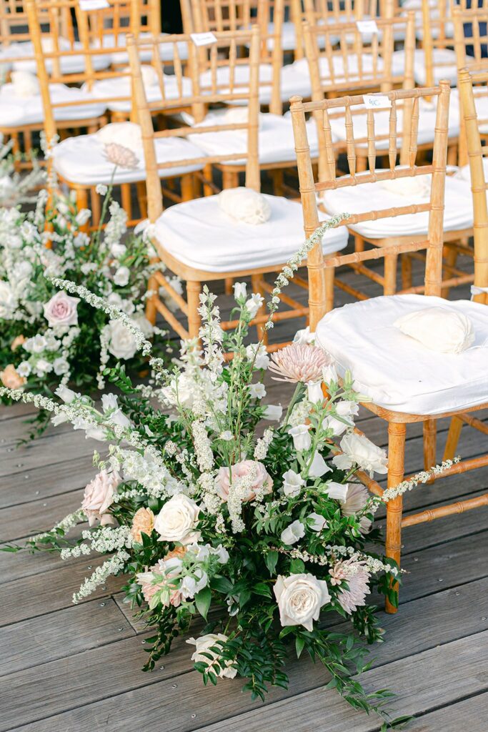 Wooden chairs with white cushions arranged in rows, adorned with white and blush floral arrangements along a wooden deck.