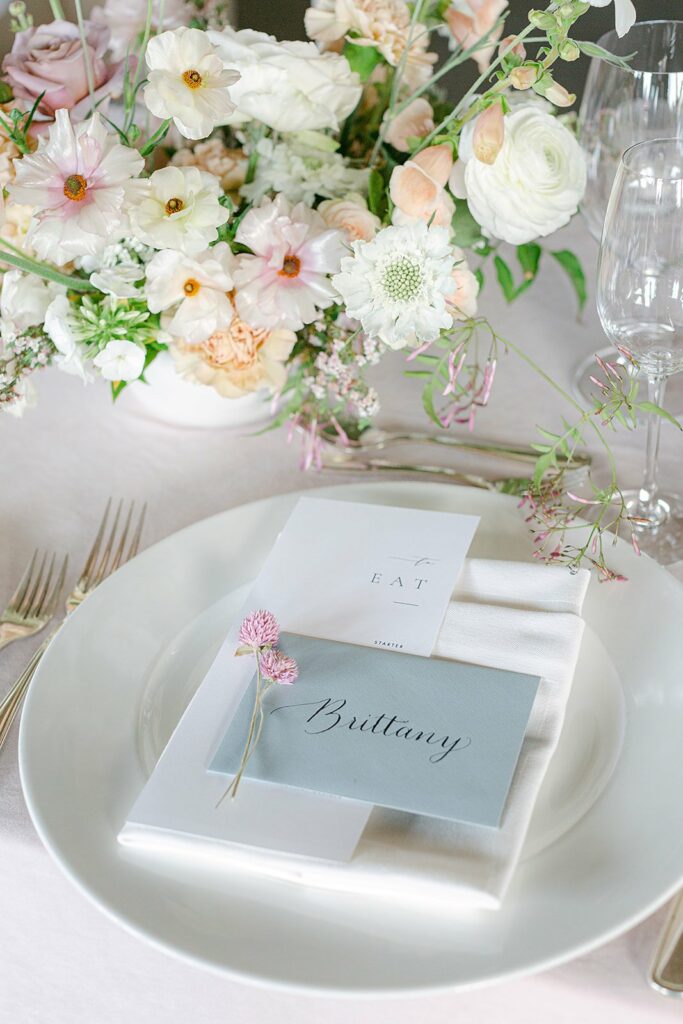Elegant table setting with floral centerpiece and a place card labeled "Brittany" on a white plate with silverware and glasses.
