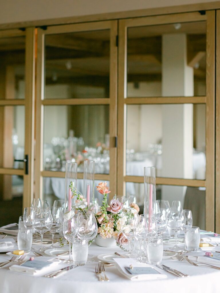 Elegantly set dining table with multiple glasses, candles, and a floral centerpiece, in front of wooden-framed glass doors in a well-lit room.