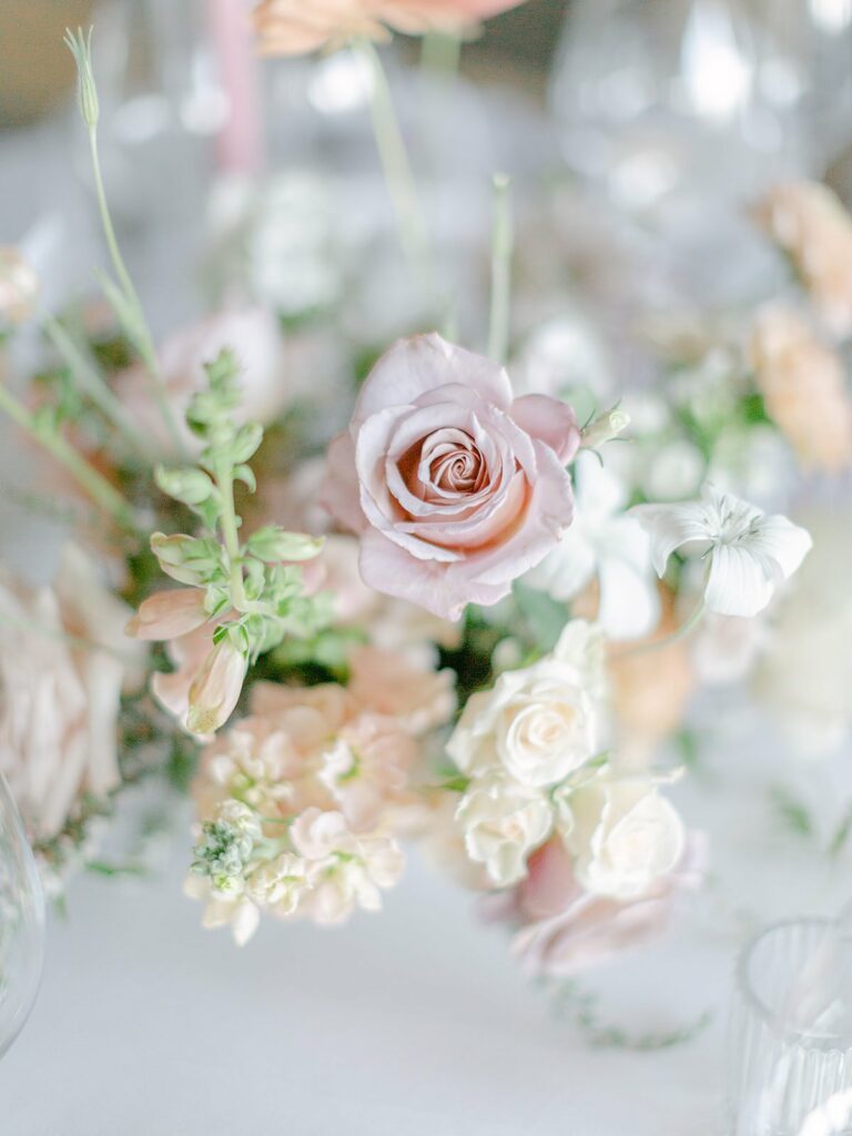 A close-up of a floral arrangement featuring light pink roses, white flowers, and green foliage, set against a blurred background.