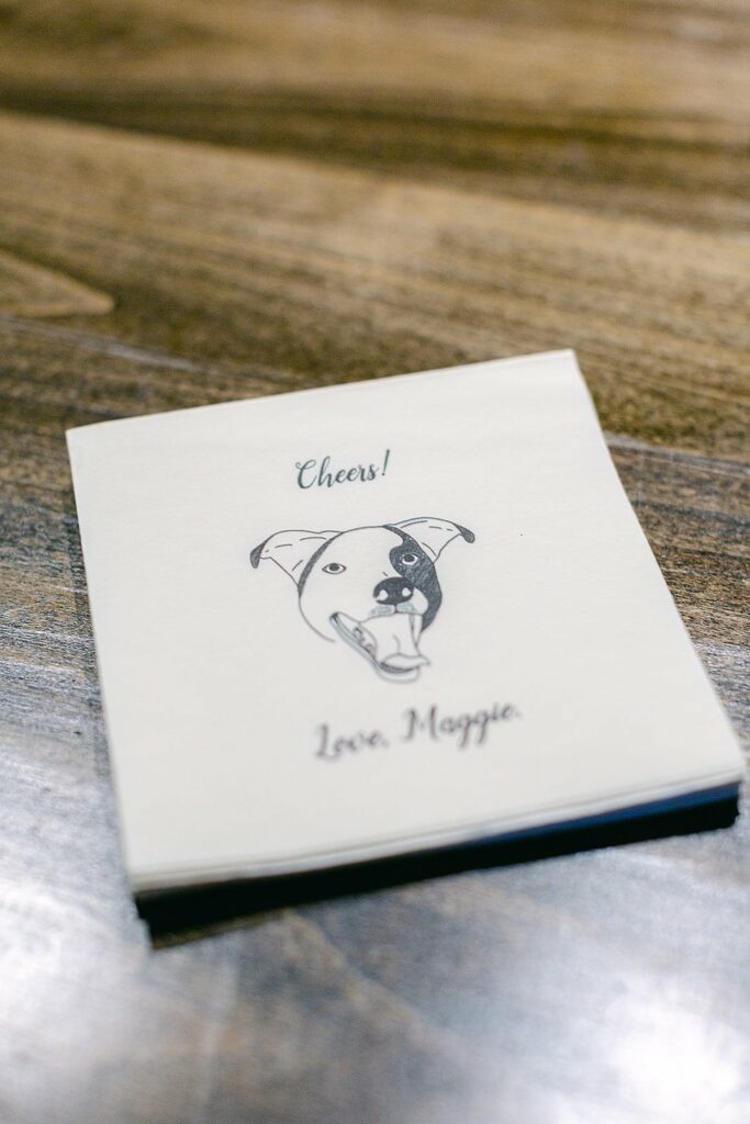 A stack of napkins on a wooden surface features an illustration of a dog and the text "Cheers! Love, Maggie.