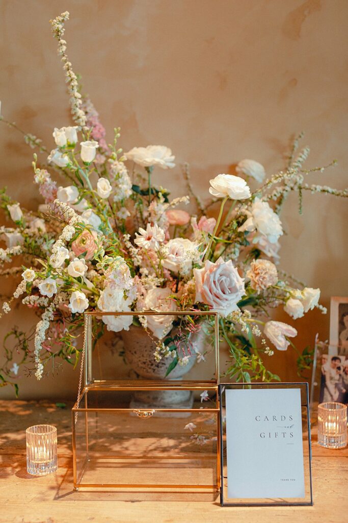 A floral arrangement in a vase is displayed behind a transparent box for cards and gifts. Two candles in glass holders are placed beside the box. A sign reads "Cards & Gifts, Thank You.