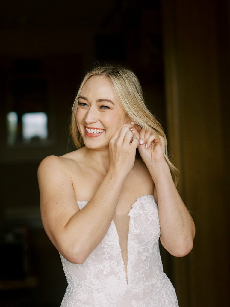 A woman in a strapless white gown smiles while adjusting her earring.
