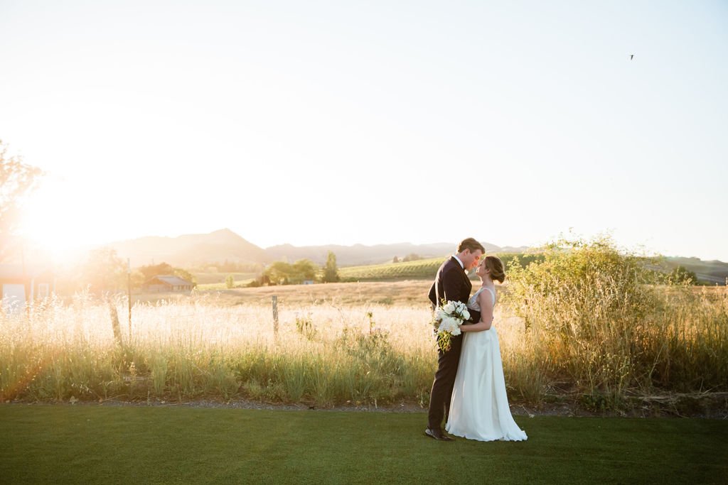A couple in wedding attire stands closely in a grassy field with the sun setting in the background, casting a warm glow over the landscape. The bride holds a bouquet of white flowers.