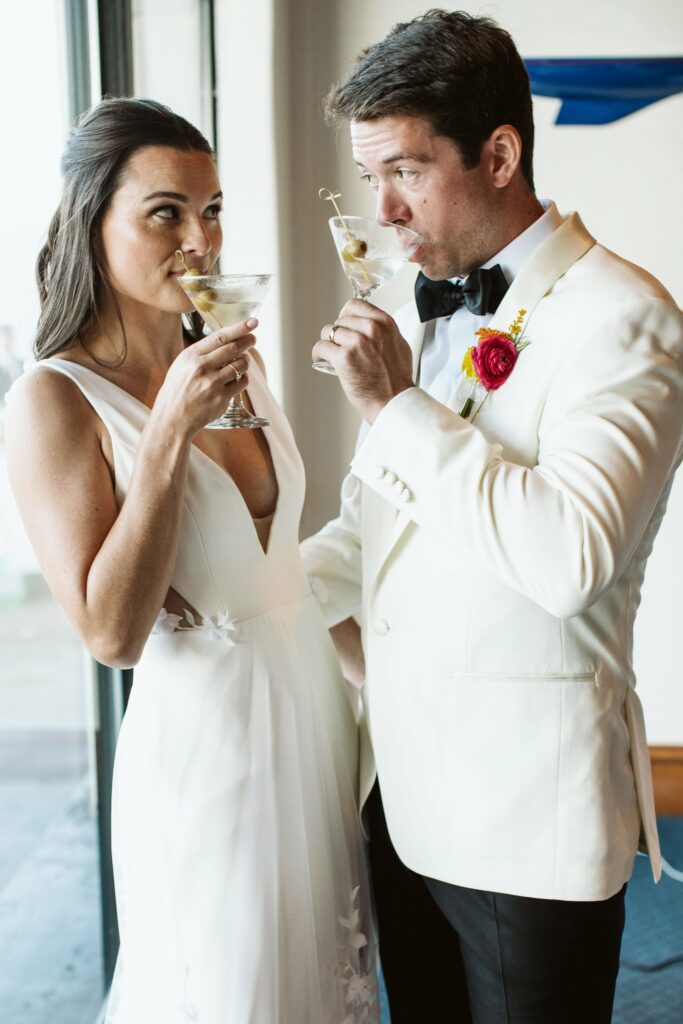 A man and a woman in formal attire share a toast with cocktails, with the man wearing a white jacket and bow tie, and the woman in a white, sleeveless dress.