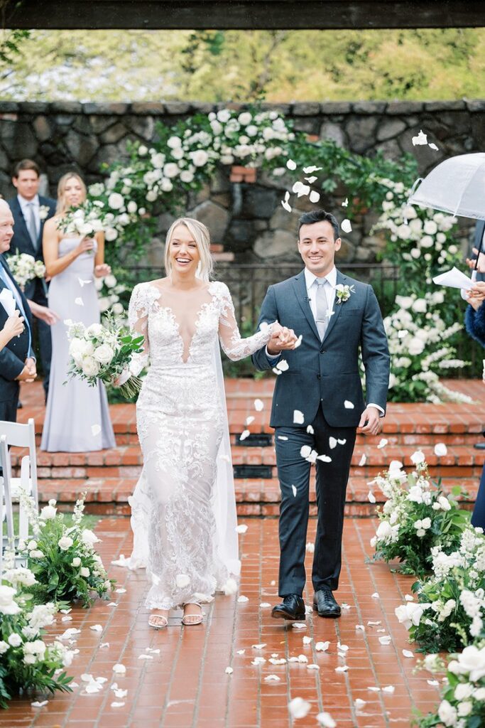 A newly married couple walks down the aisle, smiling, with flower petals being thrown. The bride wears a white lace gown and holds a bouquet; the groom wears a dark suit. Wedding guests are visible.