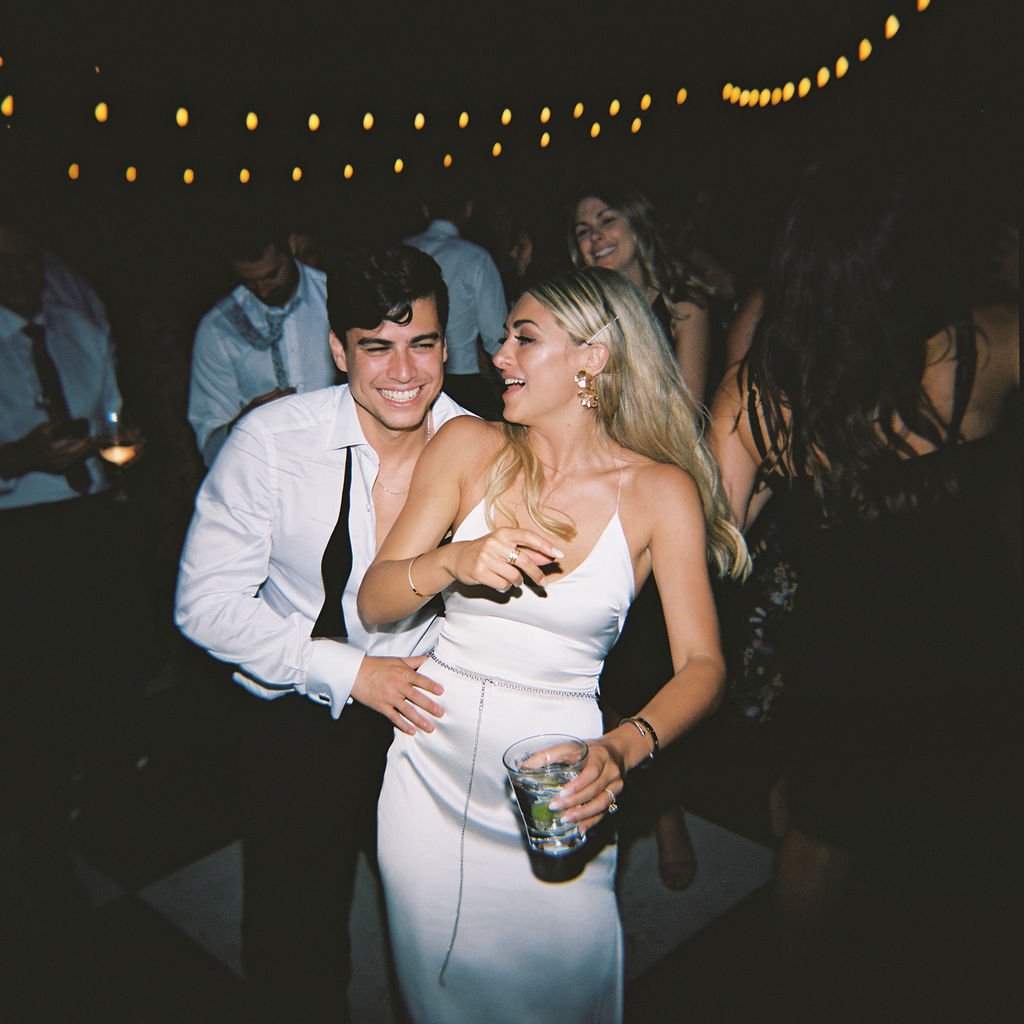 A man and woman smile and dance together at a nighttime event. string lights are visible in the background, and the woman holds a drink.