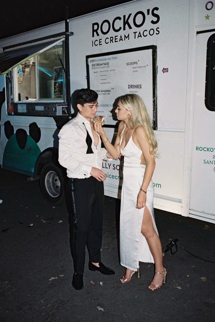 A person in a white dress holds the hand of another person wearing a white shirt and black pants in front of an ice cream taco food truck labeled "Rocko's.