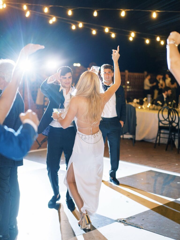 A couple dances under string lights at night, with others joining the celebration around them on a checkered floor. The woman in a white dress has her back to the camera, while the man faces it.