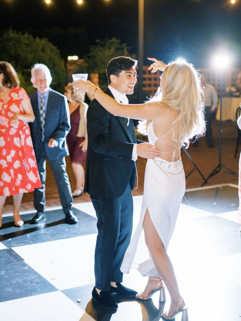 A man and woman in formal attire dance on a checkerboard floor at an outdoor evening event, while others watch and smile in the background.