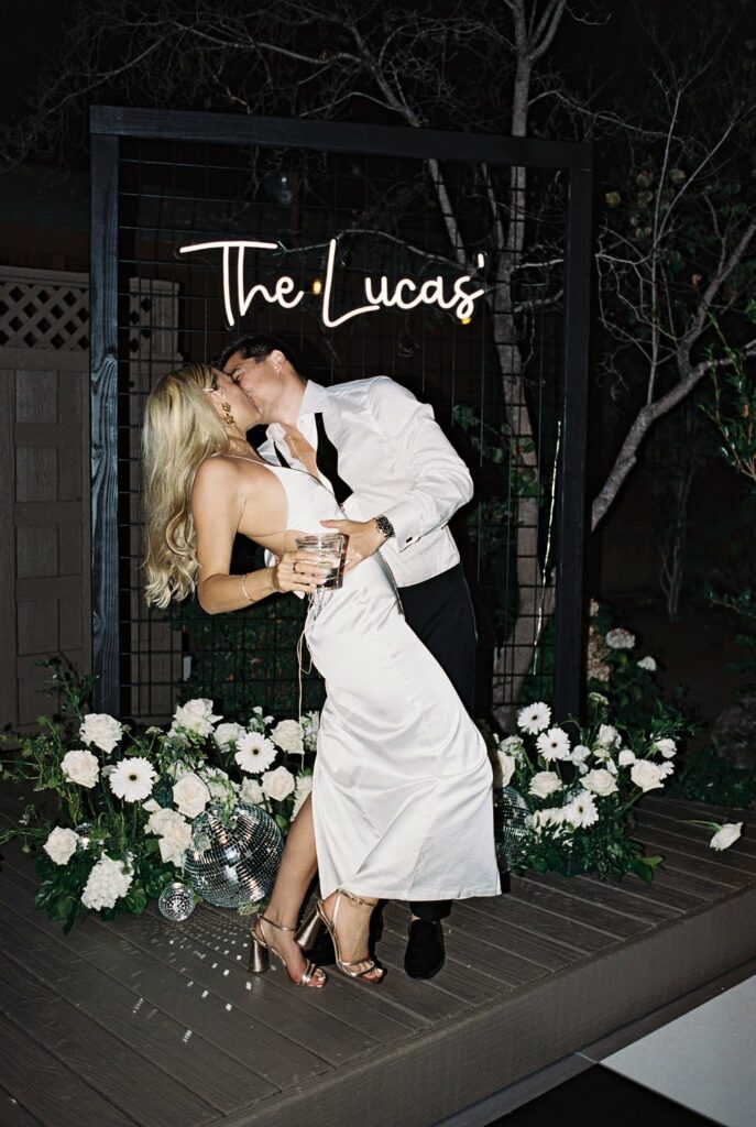 A couple dressed in formal attire share a kiss under a sign that reads "The Lucas'" surrounded by flowers and greenery at night.