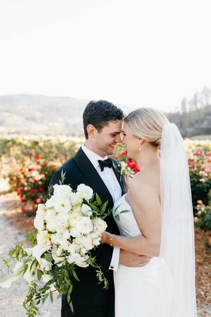 A bride and groom stand close, touching foreheads and smiling. The groom wears a black tuxedo, and the bride wears a white wedding dress and veil, holding a bouquet of white flowers. They are outdoors.
