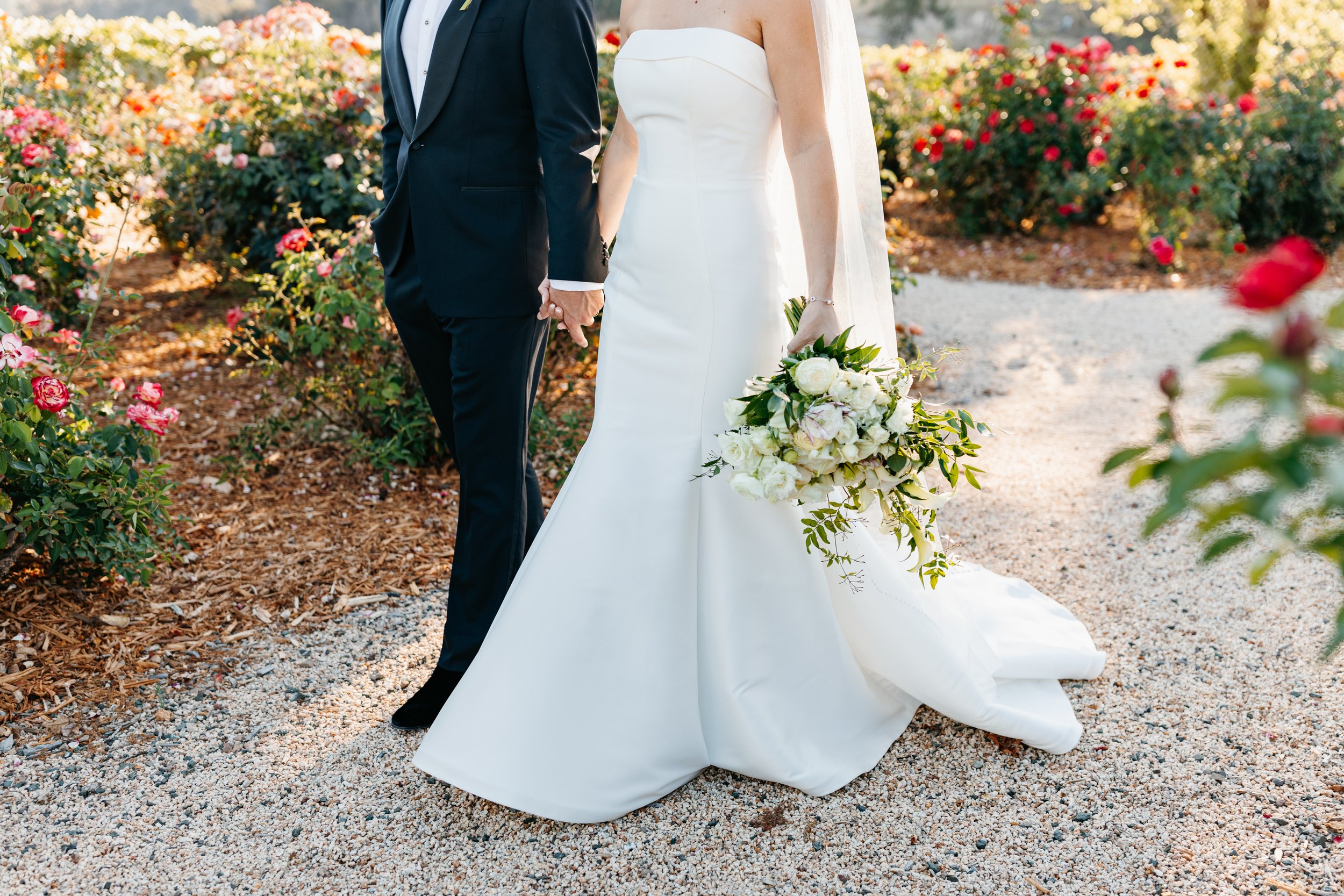 A bride in a strapless white gown holds a bouquet of white flowers while standing next to a groom in a black tuxedo, both holding hands in a garden with blooming pink and red flowers.