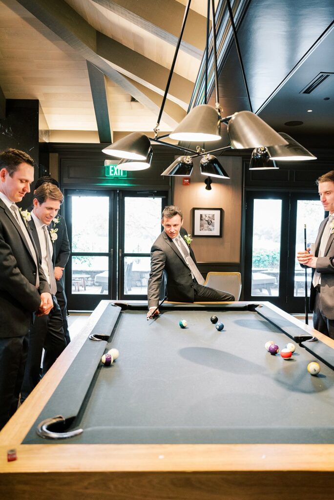 Four men in suits play pool in a well-lit room with large windows. One man is taking a shot while the others watch attentively.