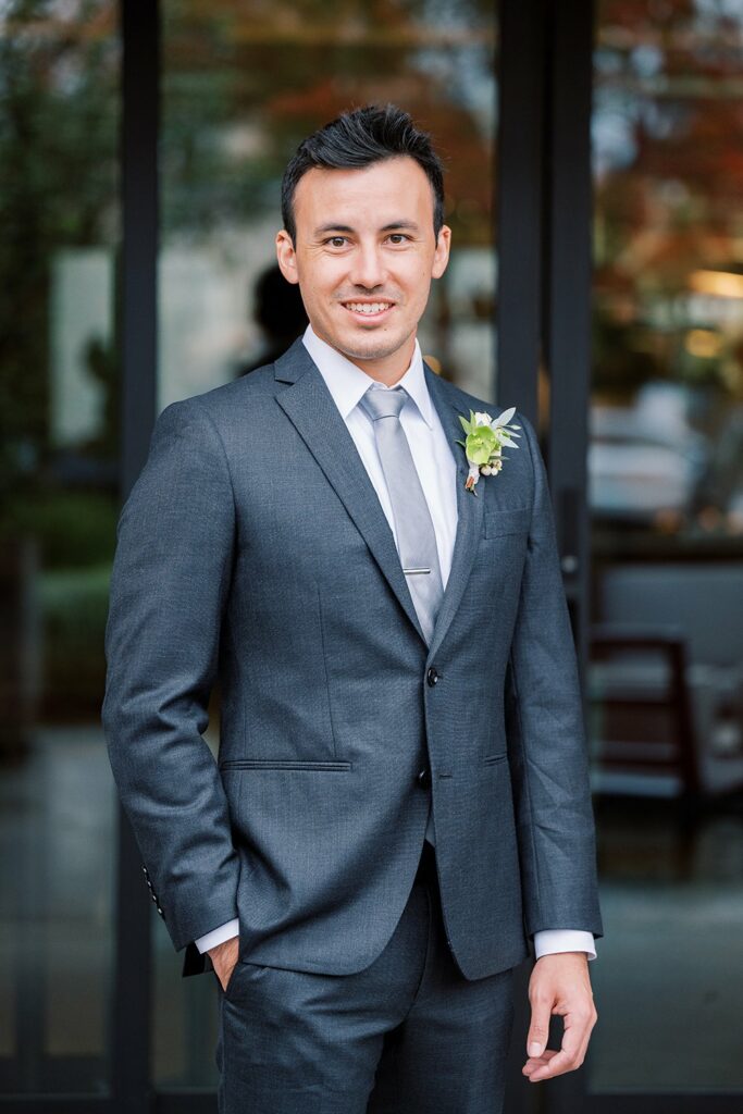 A man in a gray suit with a boutonniere stands in front of a building, posing for a formal photo with one hand in his pocket.