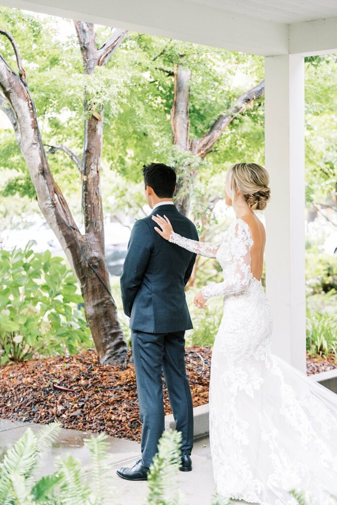 A bride in a white lace wedding dress stands behind a groom in a suit, reaching out to touch his shoulder. They are outside by a tree.