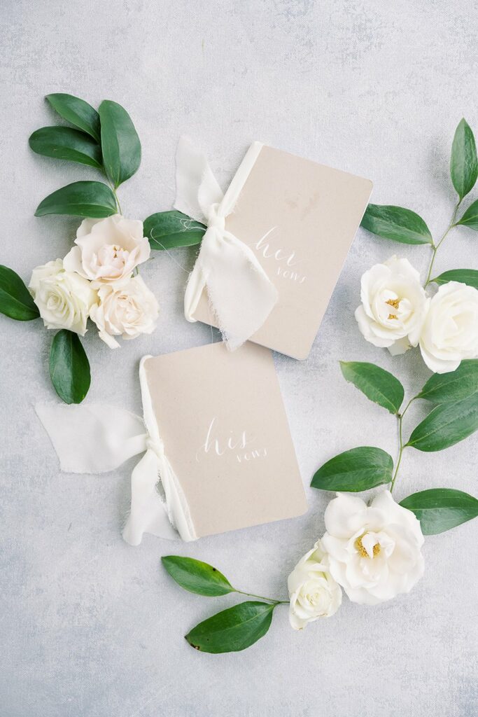 Two beige vow books labeled "his" and "hers" are placed among green leaves and white roses on a light gray surface.