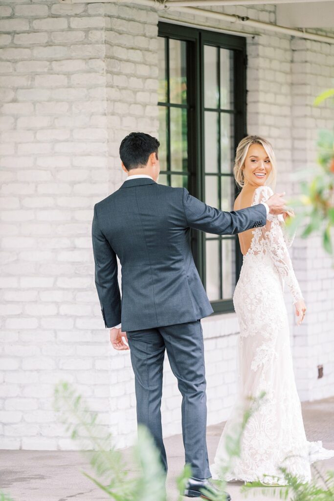 A man in a dark suit is turning a woman in a white wedding gown by her hand in front of a white brick building with large windows. The woman is smiling and looking at the camera.