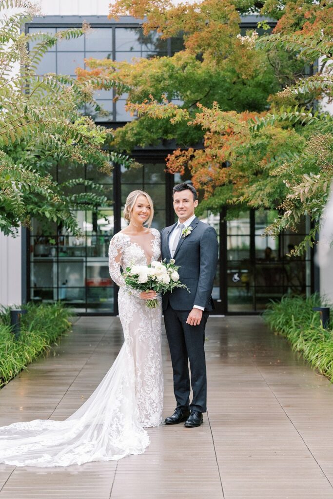 A newlywed couple stands together outdoors. The bride wears a white lace gown while the groom is in a black suit. They are both holding a bouquet of flowers and posing for a photograph.