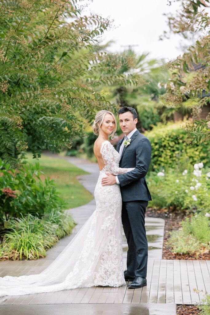A couple in wedding attire stand outdoors on a wooden path, surrounded by greenery. The bride wears a lacy white gown, and the groom is in a dark suit. They pose facing the camera, with the groom's arm around the bride.