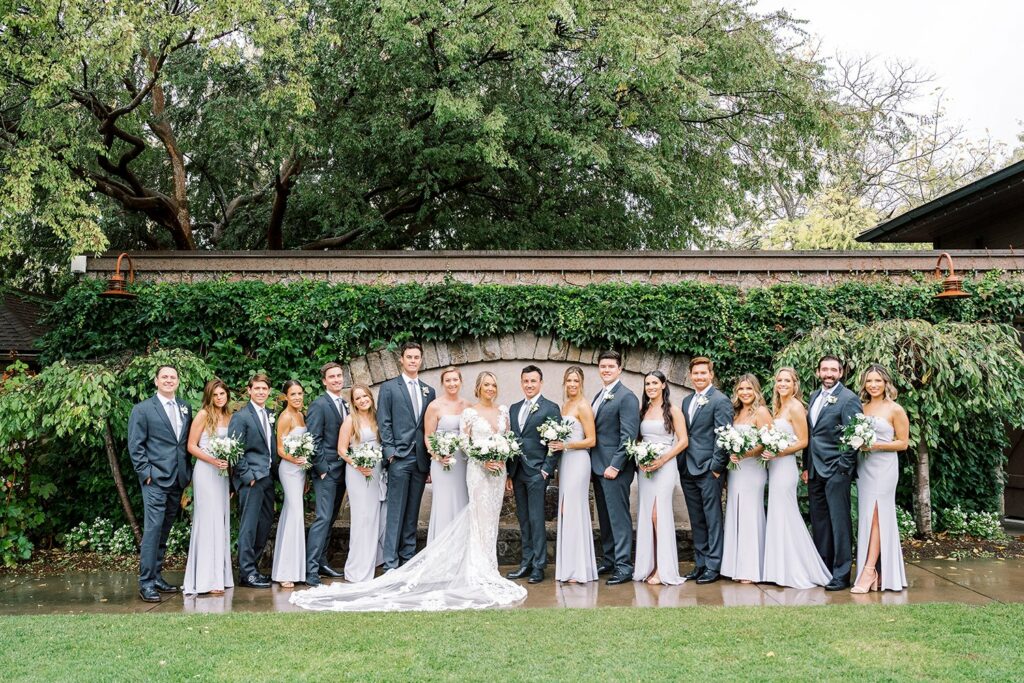 A large wedding party of bridesmaids in light gray dresses and groomsmen in dark suits stands together outdoors in front of a stone structure covered in greenery.