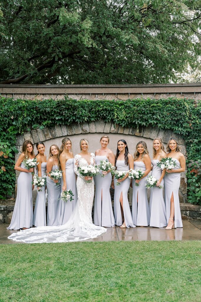 A bride in a white dress stands with nine bridesmaids in lavender dresses, each holding bouquets, in front of a stone wall covered with ivy.