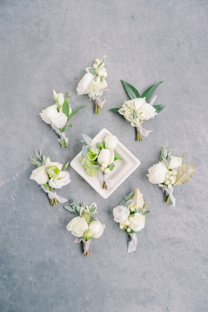 A selection of white flower boutonnieres arranged in a circle on a grey surface, with one boutonniere placed in a small white square dish in the center.