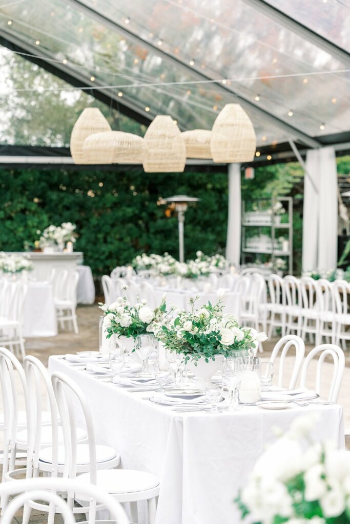 An elegant outdoor wedding reception setup with white tables, floral centerpieces, white chairs, and a transparent tent adorned with string lights and wicker lanterns.