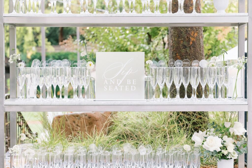 Rows of champagne glasses arranged on a metal shelf with a sign that reads "Sip and Be Seated" amidst greenery and white flowers.