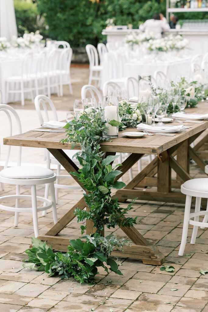 A wooden table with greenery and white candle decorations, set with plates and glasses, surrounded by white chairs on a stone patio.