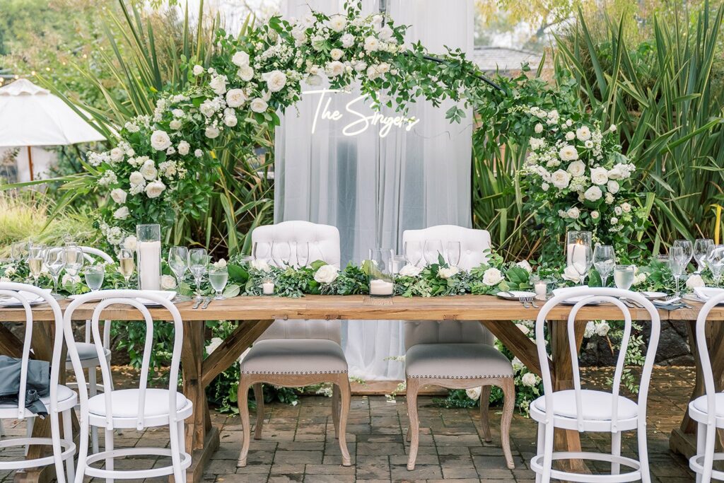 Elegantly decorated outdoor wedding table with white chairs, a wooden table adorned with greenery and white roses, and a floral arch featuring a neon "The Singers" sign in the background.