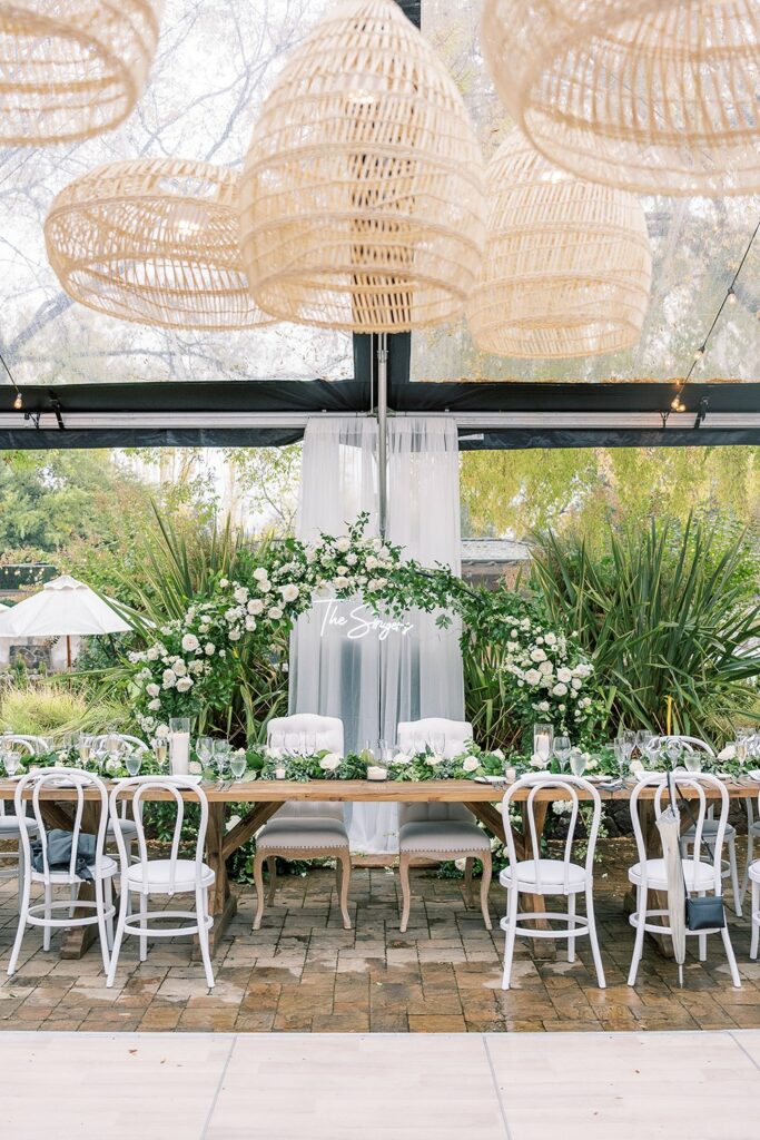 A decorated wedding reception table with white chairs, greenery, and floral arrangements, set under hanging wicker lamps with transparent roofing showing trees and the outdoors in the background.