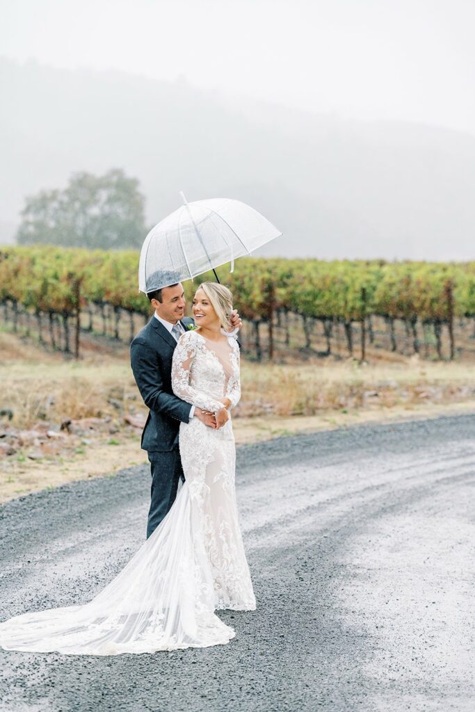 A couple in wedding attire stands on a wet road, with the groom holding a transparent umbrella over them. Vineyard rows and mist-covered hills are in the background.