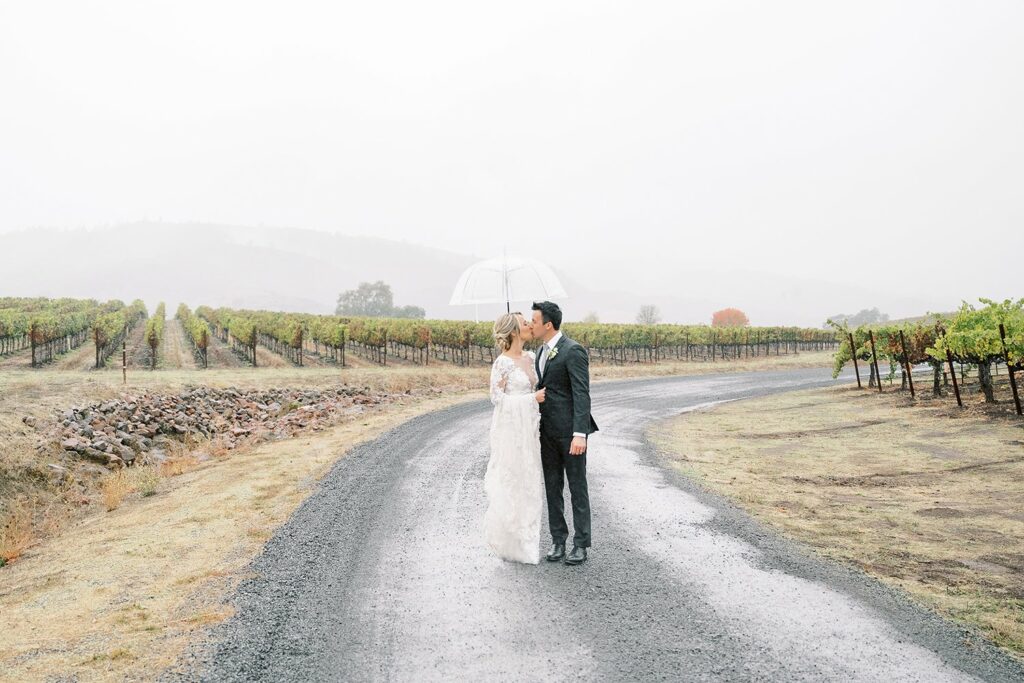 A couple stands on a gravel path in a vineyard, sharing an umbrella and a kiss in misty weather. The vineyard stretches out into the foggy distance.