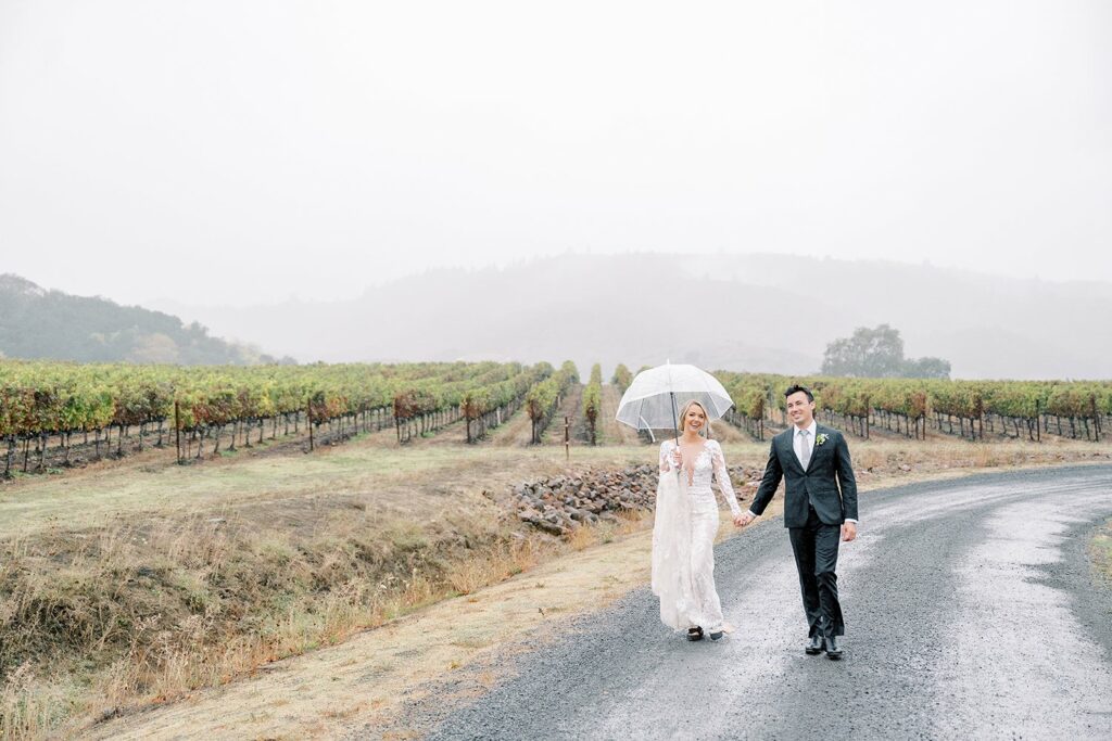 A bride and groom walk hand-in-hand on a rainy day along a vineyard road. The bride holds a clear umbrella, and both wear formal attire. The background features rows of grapevines and misty hills.