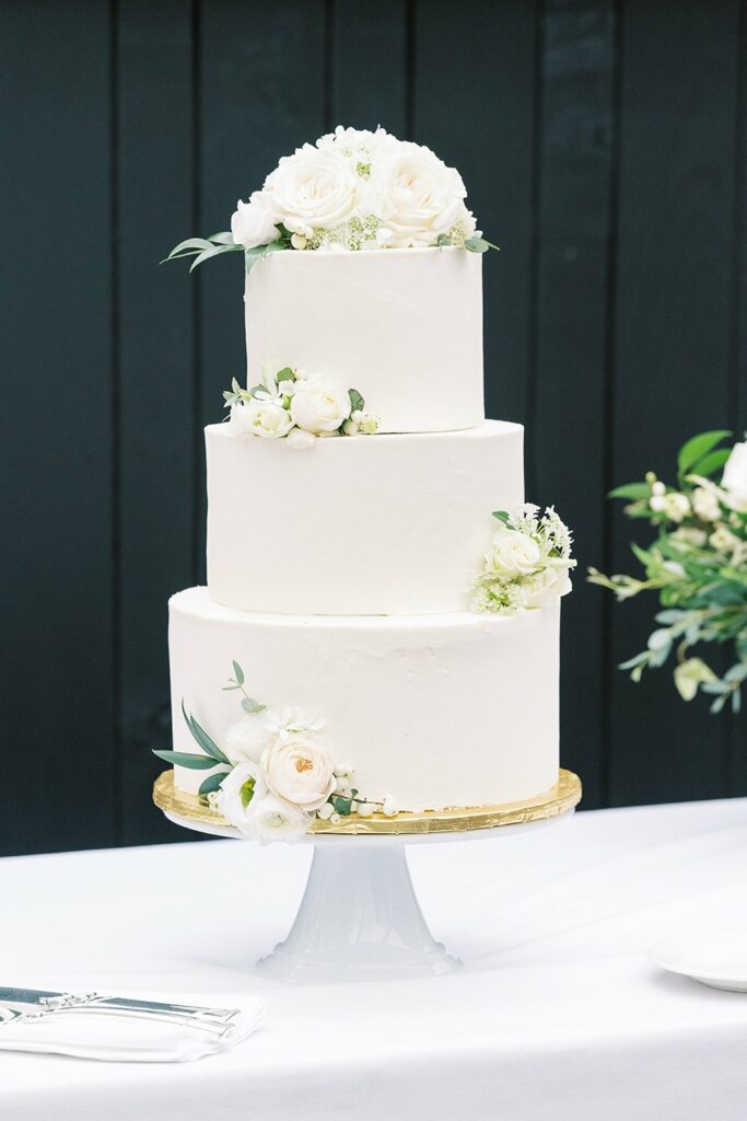 A three-tier white wedding cake adorned with white roses and greenery, placed on a white cake stand with gold trim on a white tablecloth.