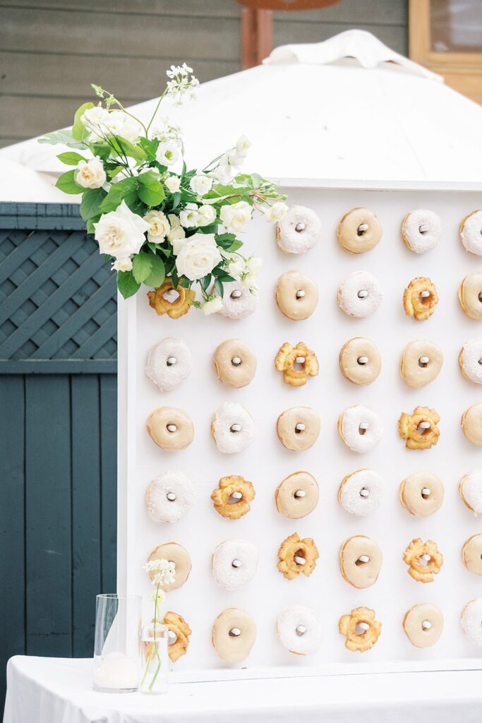 A display board is filled with various donuts arranged in even rows and columns. The top corner of the board is adorned with a small bouquet of white flowers.