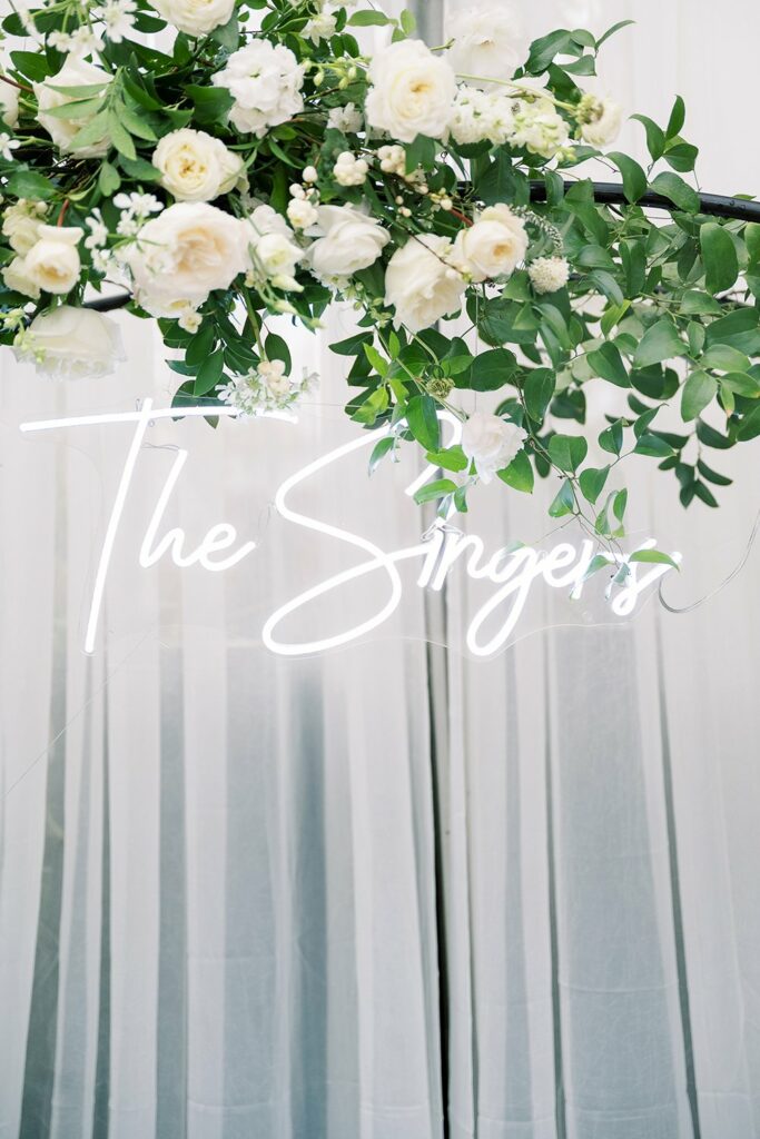 A white neon sign reading "The Singers" hangs among white flowers and green leaves in front of a white curtain background.