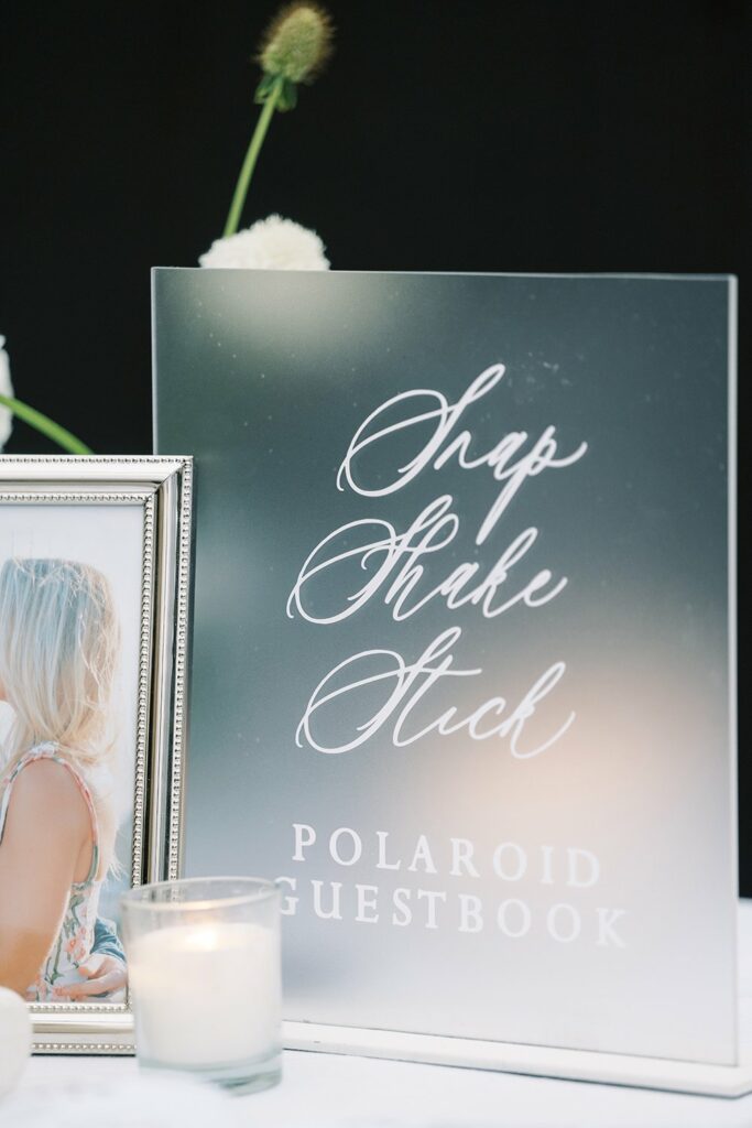Clear guestbook sign with "Snap, Shake, Stick" instructions in elegant script, next to a framed photo and a lit candle.