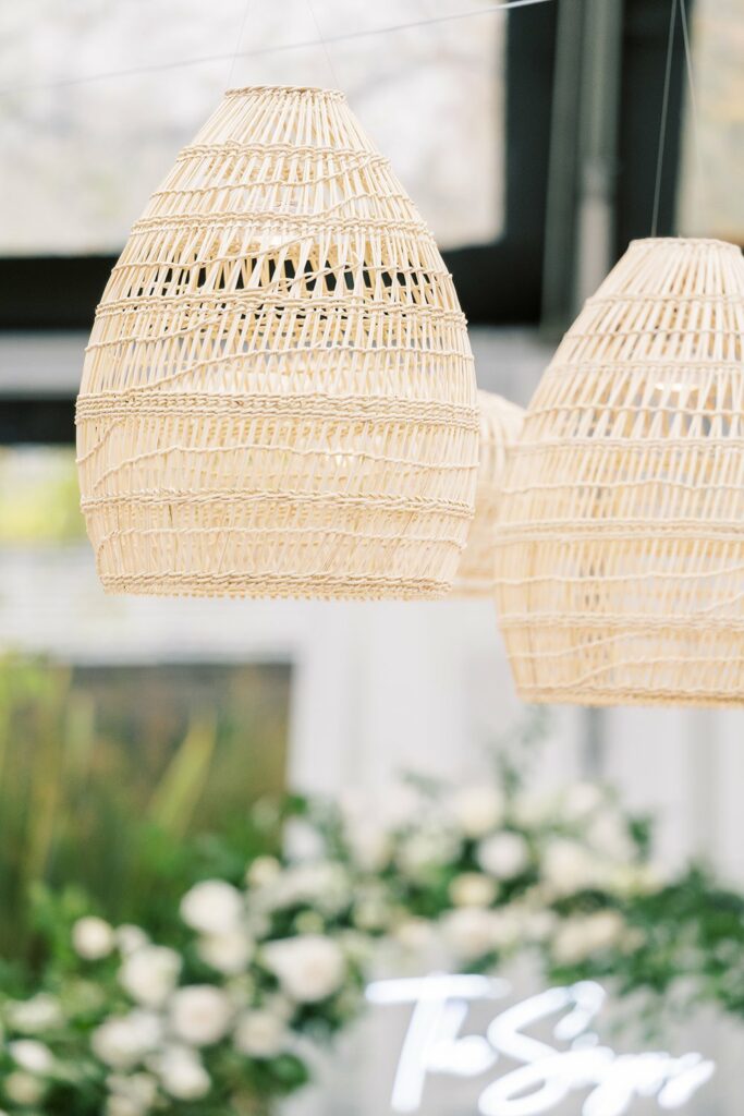 Close-up of woven wicker light fixtures hanging from the ceiling, with a blurred background featuring greenery and white flowers.