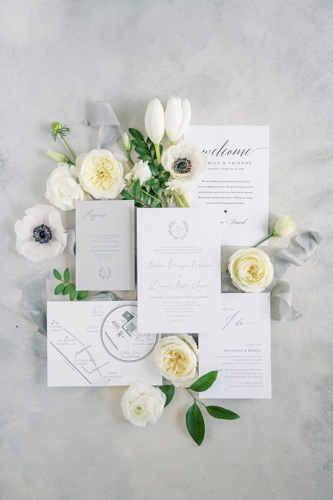 A wedding invitation suite is displayed on a light surface, featuring white and cream floral arrangements and delicate greenery. A map and various invitation cards are visible among the decor.