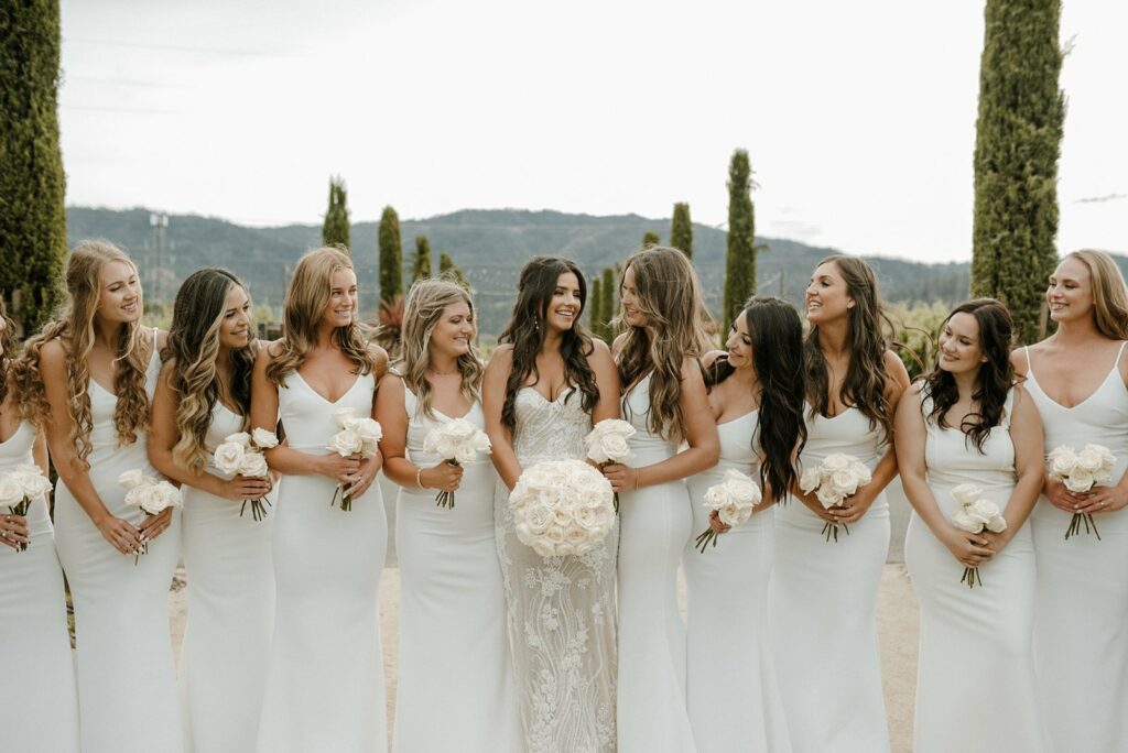 A bride and ten bridesmaids in white dresses stand in a row outdoors, each holding a bouquet of white roses. Tall, narrow trees are in the background.