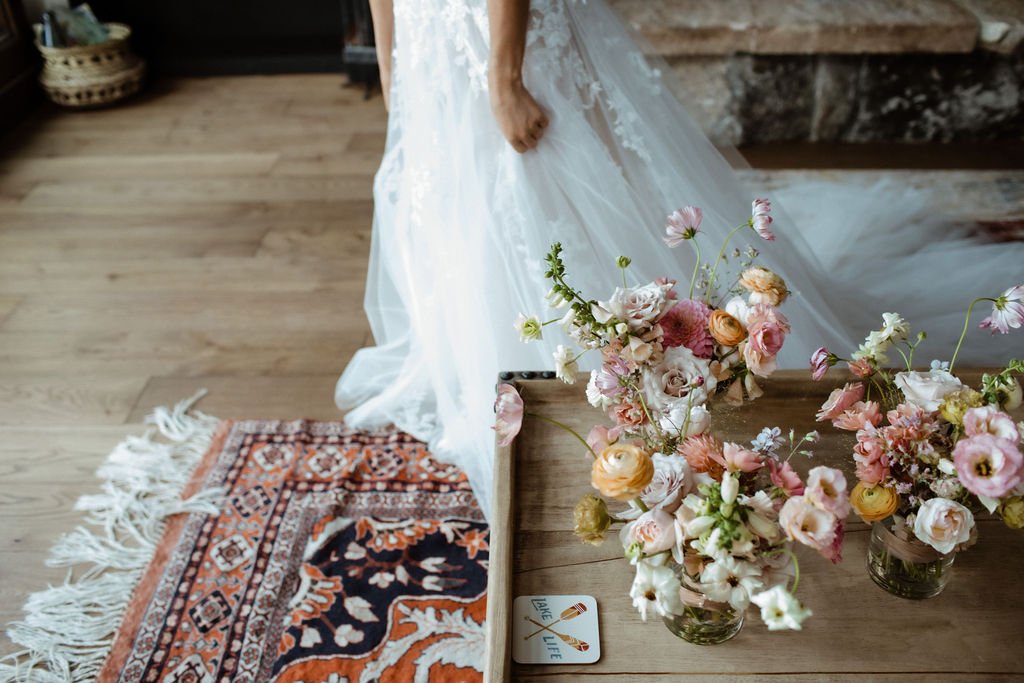 A person in a white dress stands beside a wooden table with bouquets of flowers and a colorful rug on the floor.