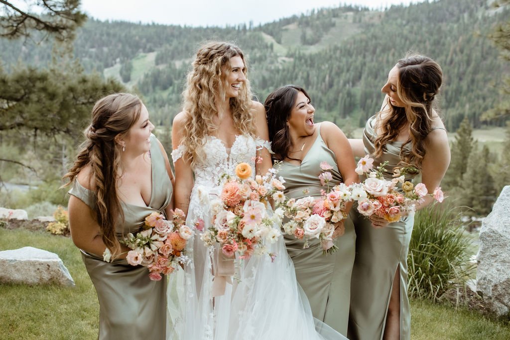A bride and three bridesmaids, all holding bouquets, smile and chat outdoors with a scenic forested mountain background.