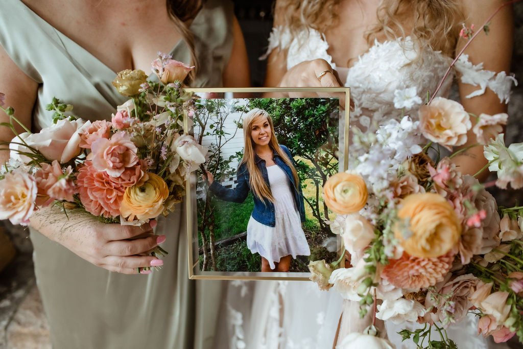 Two women in dresses hold a framed photo of another woman and bouquets of flowers.