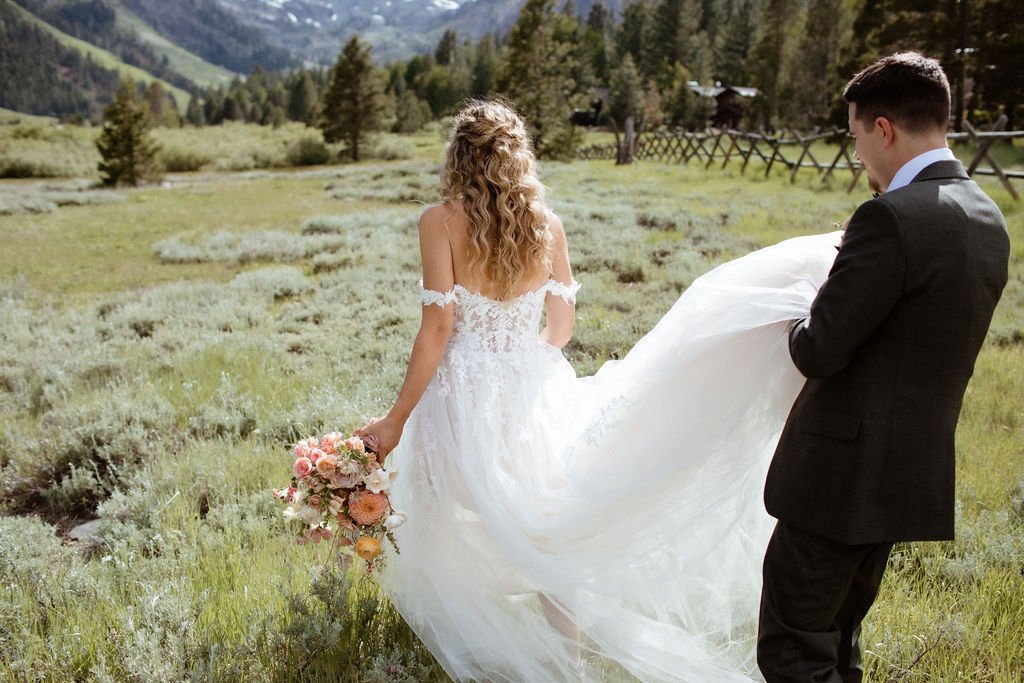 A bride in a white dress holding a bouquet and a groom in a suit stand in a grassy field with trees and mountains in the background.