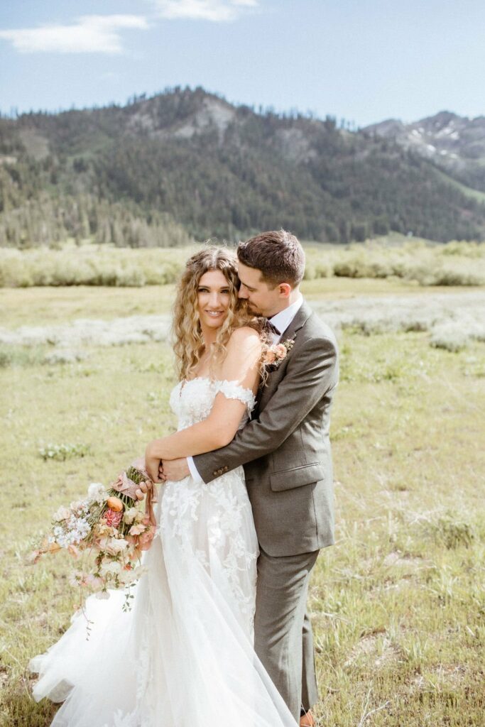 A bride and groom stand together in a field with mountains in the background. The bride in a white dress holds a bouquet, and the groom in a gray suit embraces her.