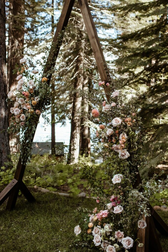 A triangular wooden wedding arch is adorned with a variety of pink, peach, and white flowers set against a backdrop of a wooded area and a body of water.